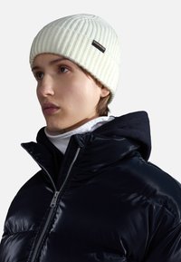 Berretto a maglia crema a costine con etichetta nera, indossato con una giacca a vento nera con chiusura a zip e interno blu navy con cappuccio.
