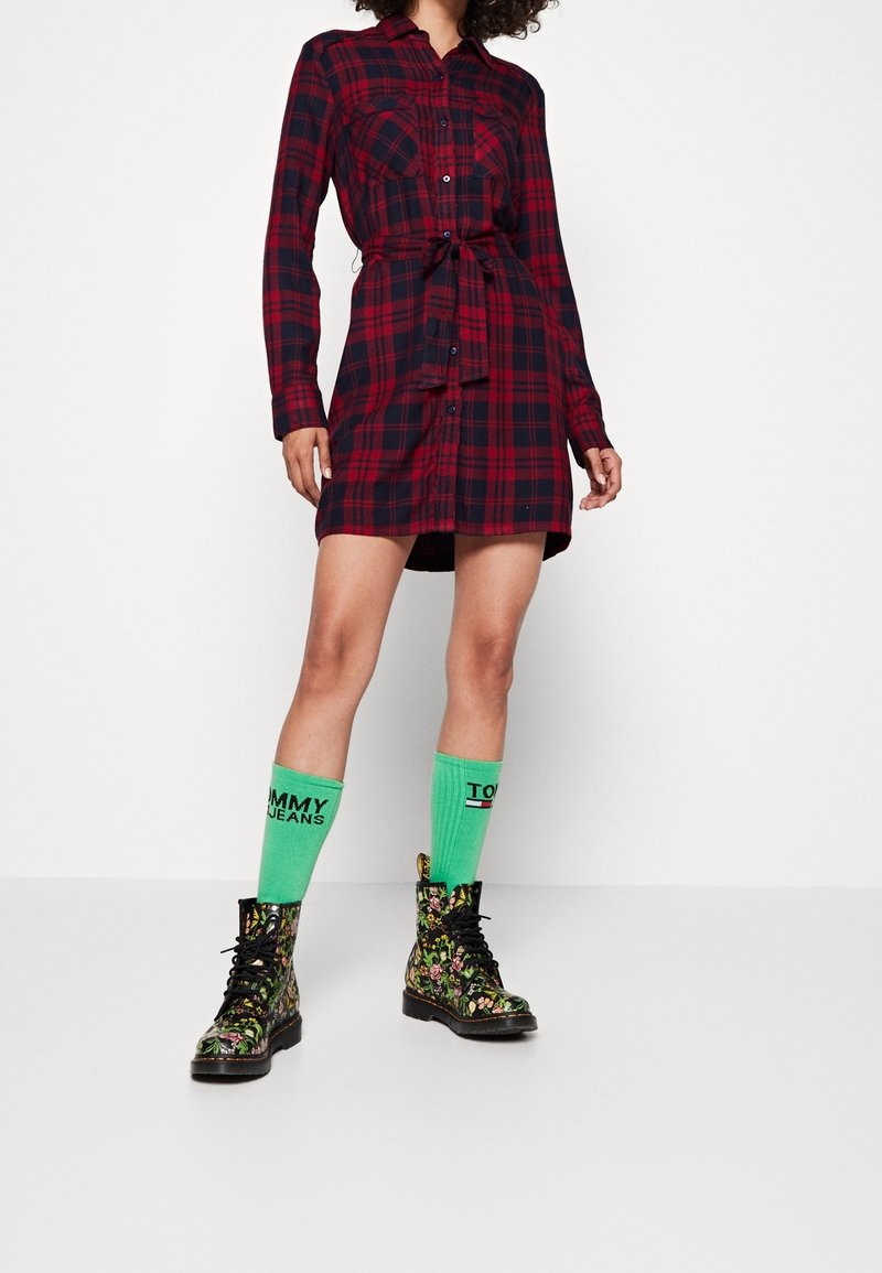 Personne portant une robe chemise à carreaux rouge et noir, des chaussettes vert vif Tommy Jeans, et des bottines noires à lacets à motif floral, debout sur un fond blanc.