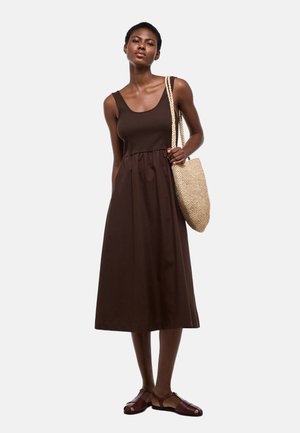 Femme portant une robe midi sans manches marron foncé, tenant un sac à bandoulière tissé beige et portant des sandales plates marron foncé.