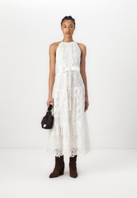 Robe maxi en dentelle blanche avec un décolleté halter, une taille cintrée et une jupe à volants ; associée à un petit sac à main noir et des bottes à talons marron.