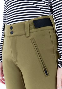 Olijfgroene rekbare broek met twee zwarte knopen, een ritsvak en een zwarte verstelbare tailleband, gecombineerd met een gestreepte lange mouw top.
