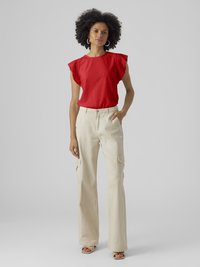 Γυναίκα που στέκεται φορώντας κόκκινη αμάνικη μπλούζα με τρύπες, μπεζ παντελόνι cargo, κρίκους στα αυτιά και σανδάλια ανοιχτής μύτης σε απλό γκρι φόντο.