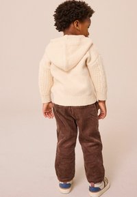 Sudadera de punto crema con mangas texturizadas, combinada con pantalones de pana marrón. Presenta una capucha y calzado casual con acentos multicolor.