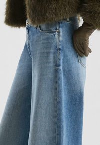 Jeansy typu wide-leg w średnim niebieskim denimu, z wysokim stanem, bocznymi kieszeniami i gładką fakturą. Akcentowane brązowymi skórzanymi rękawiczkami.