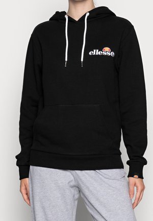 Personne portant un sweat à capuche noir ellesse avec des cordons blancs et un pantalon gris clair, debout devant un fond uni.