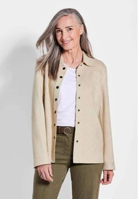 Veste en daim beige avec un col, fermée par des boutons à l'avant, et des manches longues, portée sur une chemise blanche et un pantalon vert olive.