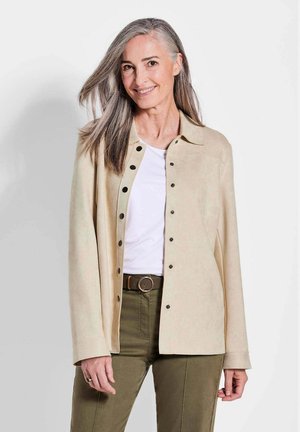 Giacca in suede beige con colletto, chiusura a bottoni sul davanti e maniche lunghe, indossata sopra una camicia bianca e pantaloni verde oliva.