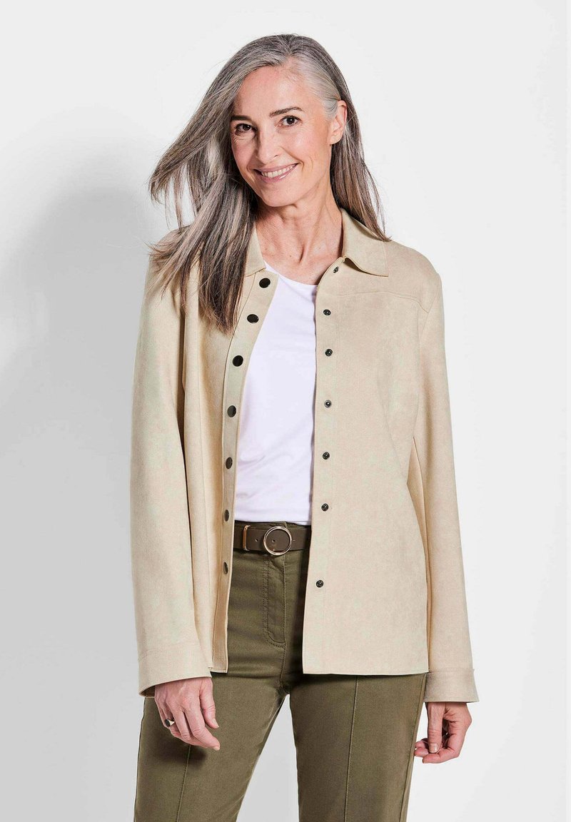 Veste en daim beige avec un col, fermée par des boutons à l'avant, et des manches longues, portée sur une chemise blanche et un pantalon vert olive.