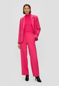 Roze op maat gemaakte blazer en wijde broek, gecombineerd met een top met hoge hals. Zachte textuur, gestructureerd ontwerp, aangevuld met puntige zwarte hakken.
