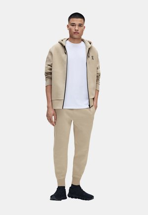 Jeune homme debout portant un sweat à capuche beige, un pantalon de jogging assorti, un T-shirt blanc et des chaussures de sport noires sur un fond clair uni.