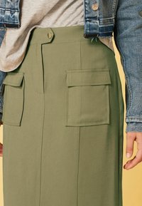 Falda cargo verde oliva con dos bolsillos frontales de parche, diseño plano en la parte delantera y cierre de botón a presión, combinada con una chaqueta de mezclilla.