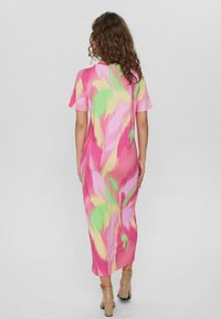 Kobieta z kręconymi włosami, ubrana w midi sukienkę w abstrakcyjny wzór w odcieniach różu, zieleni i żółci oraz beżowe sandały na wysokim obcasie, zwrócona plecami.