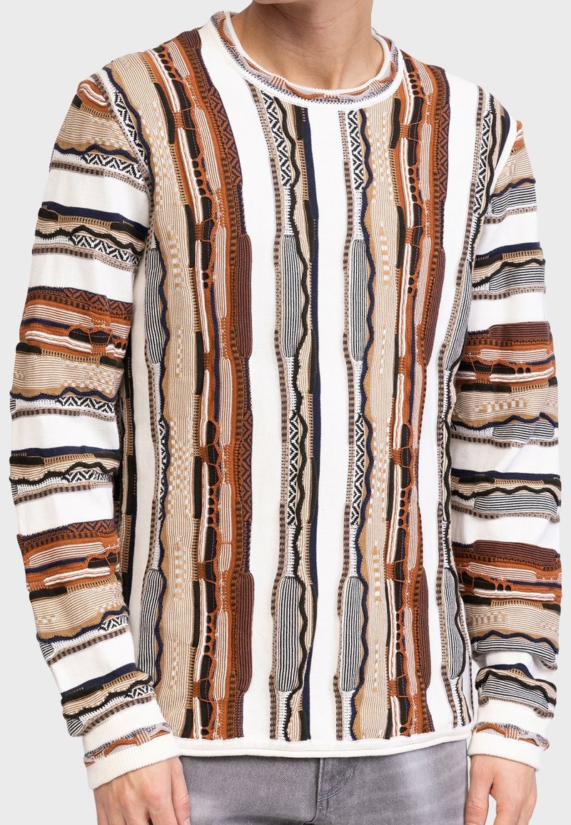 Gros plan sur une personne portant un pull à manches longues avec des rayures texturées verticales en tons blanc, marron, noir et beige.