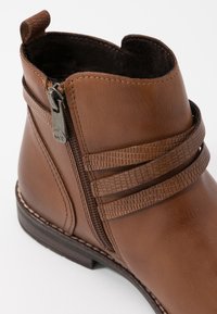 Braunes Leder-Ankle-Boot mit einem strukturierten Riemen, seitlichem Reißverschluss und genähtem Absatz. Das Innere ist mit dunklem Stoff gefüttert für zusätzlichen Komfort.