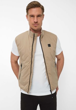 Mann mit kurzen blonden Haaren, der eine beige ärmellose Jacke mit Reißverschluss über einem weißen T-Shirt trägt, steht mit den Händen in den Taschen vor einem weißen Hintergrund.