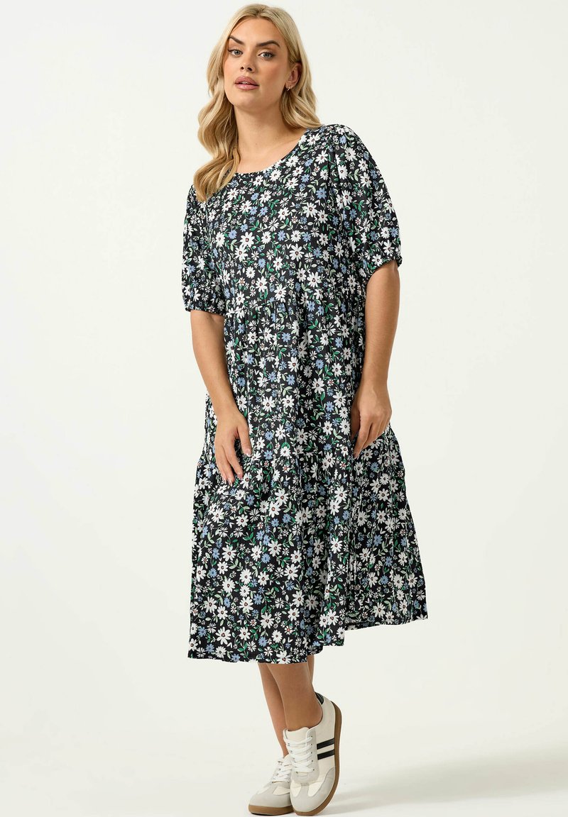 Frau trägt ein schwarzes Midikleid mit weißem und blauem Blumenmuster, Puffärmeln und weißen Sneakers mit schwarzen Streifen.