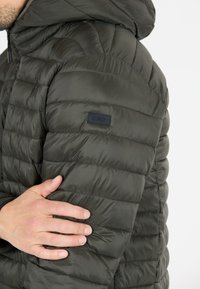 Mörkgrön puffjacka med huva, med kviltade sektioner, slät nylontextur och en liten svart logotyp på ärmen.