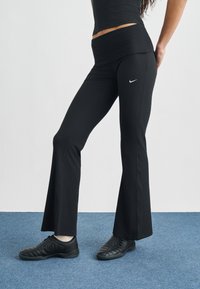 Μαύρο φαρδύ παντελόνι γιόγκα με ριμπ υφή και φαρδιά ζώνη μέσης, με μικρό λευκό λογότυπο Nike στο ισχίο. Φορεμένο με μαύρα αθλητικά παπούτσια.