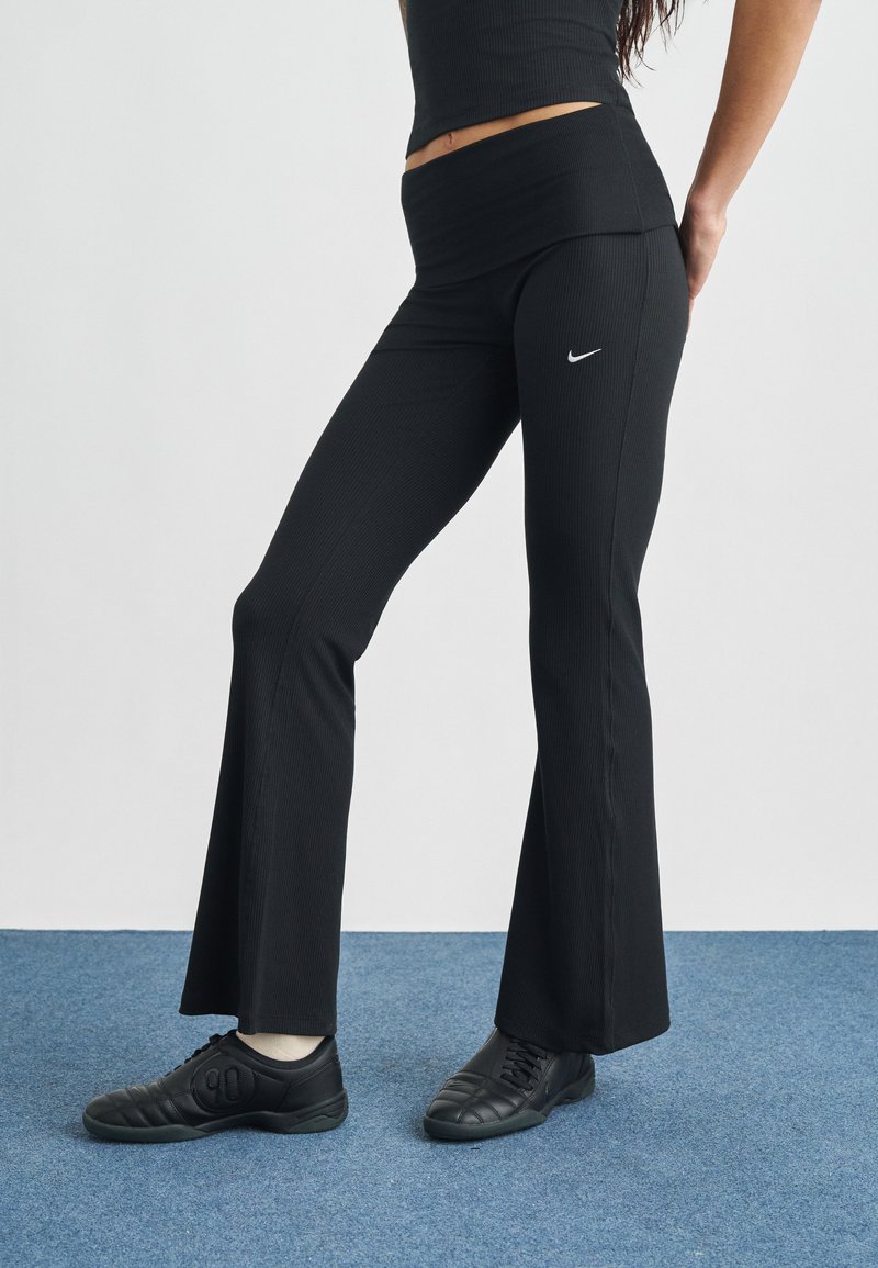 Μαύρο φαρδύ παντελόνι γιόγκα με ριμπ υφή και φαρδιά ζώνη μέσης, με μικρό λευκό λογότυπο Nike στο ισχίο. Φορεμένο με μαύρα αθλητικά παπούτσια.