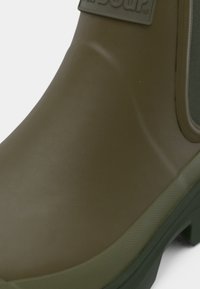 Oliwkowo zielony gumowy kalosz o gładkiej powierzchni, z zaokrąglonym noskiem i teksturowaną podeszwą, wyposażony w pętlę do zakładania i logo po boku.