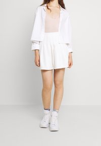 Bílá košile, světle růžový body a bílé šortky s vysokým pasem. Modelka v bílých teniskách a ponožkách s batikováním. Hladké textury, ležérní styl.