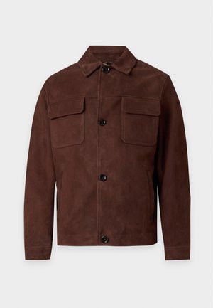 Uomo che indossa una giacca marrone sopra una camicia bianca, pantaloni marrone scuro e scarpe marroni, in piedi davanti a uno sfondo chiaro e semplice.
