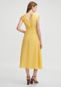 Żółta lniana sukienka z dekoltem w kształcie litery V oraz z tyłu, sięgająca do połowy łydki, z dopasowaną talią. Połączona z brązowymi sandałami na obcasie z paskami.