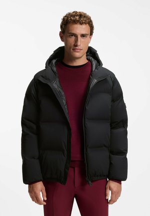 Mann trägt eine schwarze Steppjacke über einem bordeauxfarbenen Pullover und passenden bordeauxfarbenen Hosen, steht vor weißem Hintergrund.