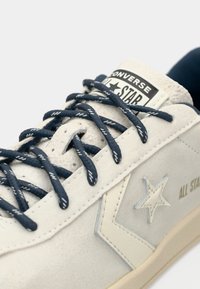 Kremfarget semsket sneaker med marineblå lisser, med stjernelogo, teksturerte detaljer og en hvit tungeetikett som viser "Converse All Star."