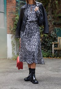 Schwarze Lederbikerjacke über einem langen, dunkel gemusterten Kleid mit floralen Motiven. Glänzende schwarze Knöchelstiefel mit dicken Sohlen und roter Handtasche.