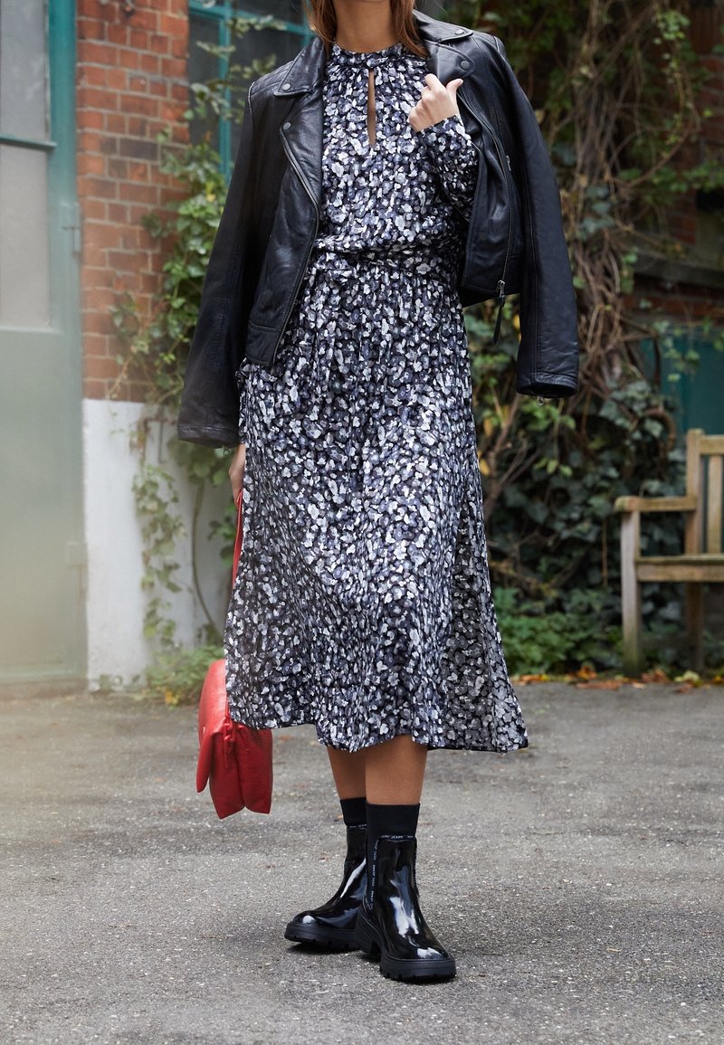 Schwarze Lederbikerjacke über einem langen, dunkel gemusterten Kleid mit floralen Motiven. Glänzende schwarze Knöchelstiefel mit dicken Sohlen und roter Handtasche.