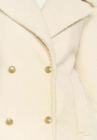 Dwurzędowy płaszcz z miękkiej, jasnobeżowej tkaniny z guzikami w kolorze złotym i klapami z wycięciami. Faktura wygląda na puszystą i ciepłą.