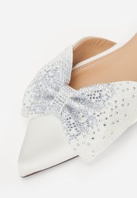 Valkoinen satiini tasapohjainen kenkä, jossa on terävä kärki ja suuri, rhinestone-koristeltu nauha edessä, jonka pinnassa on tekstuuripinta.