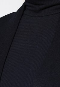 Maglione nero a collo alto a coste, caratterizzato da un design aderente e testurizzato con un colletto liscio e un tessuto morbido, parzialmente visibile nell'immagine.