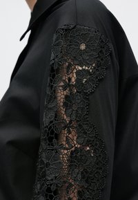 Fekete blúz, amely bonyolult fekete virágmintás csipkedíszítéssel rendelkezik a bal ujjon, kiemelve a sima anyagot és az elegáns gallér kialakítást.