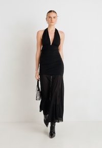 Rochie neagră cu decolteu halter, cu textură drapată, suprapusă cu un voal lung până la podea și siluetă mulată. Completată de o mică geantă argintie.