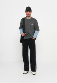 Felpa grigia con logo rosa, maniche blu, pantaloni oversized neri e calzature bianche. Un cappellino nero completa il look casual.