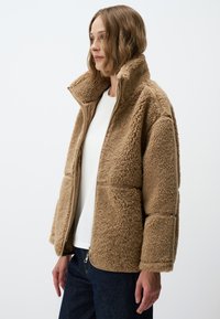 Beige fleecejack met hoge kraag, rits sluiting en patchzakken. Heeft een gestructureerd oppervlak en gladde stofaccenten.