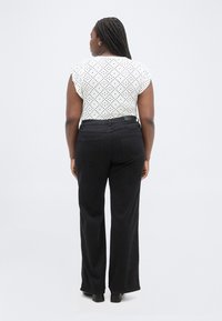 Γυναίκα με πλεξούδες στέκεται με την πλάτη, φοράει λευκό κοντομάνικο τοπ με σχέδια, μαύρο παντελόνι ψηλόμεσο με φαρδιά μπατζάκια και μαύρα παπούτσια.