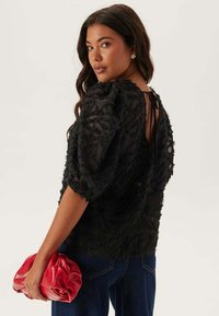 Top nero trasparente con design floreale testurizzato, maniche a sbuffo, schiena aperta con stringa, abbinato a una pochette plissettata rossa e jeans blu.