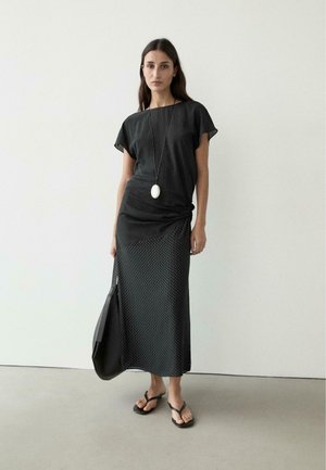 Donna con top nero testurizzato, gonna lunga con nodo laterale, infradito nere, borsa nera e una lunga collana con ciondolo bianco.