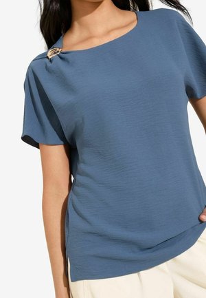 Vrouw draagt een blauw gestructureerd shirt met korte mouwen en een gouden decoratieve gesp op de linker schouder, en lichte broek.