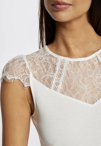 White top with a lace shoulder design featuring delicate floral patterns and scalloped edges; smooth texture on the body section.