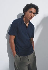 Temno modra polo majica iz mehke tkanine s kratkimi rokavi in ovratnikom, kombinirana s svetlo sivimi hlačami. Model pozira ob beli steni.