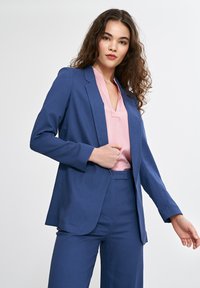 Niebieska marynarka dopasowana, wykonana z teksturowanej tkaniny, z pojedynczym zapięciem na guzik i wyraźnie podkreślonymi ramionami, zestawiona z różową bluzką pod spodem.
