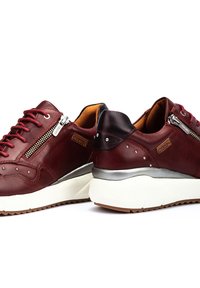 Zapatillas de cuero burdeos con acentos plateados, cremalleras laterales texturizadas, detalles de tachuelas y suela de goma blanca. Diseño de tacón ligeramente elevado.