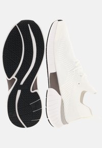 Chaussures de sport en maille blanche avec une semelle en caoutchouc noire présentant un motif antidérapant texturé. Système de laçage intégré et accents marron clair.