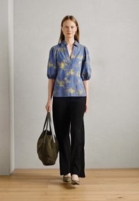 Svetlo modra bluzica z rumenim vzorcem, napihnjenimi rokavi in ostrim ovratnikom, v kombinaciji s črnimi hlačami in zeleno torbico, na lesenem tleh.