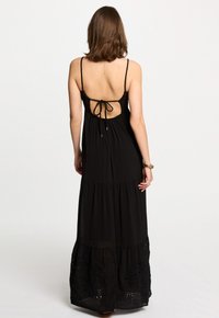 Robe maxi noire à fines bretelles, design dos nu et jupe à volants avec des détails brodés au bord. Tissu doux et fluide.