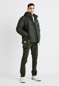 Giacca verde imbottita con cappuccio, chiusura con zip e logo. Pantaloni in corduroy verde oliva, borsa a tracolla nera e sneakers bianche con dettagli turchesi.